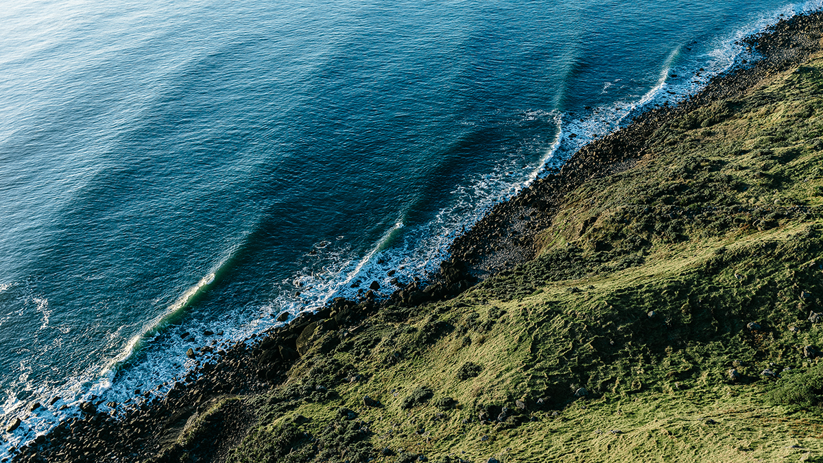Birds eye view of a rugged New Zealand coast line, sea, a rocky cliff and a grassy hill.